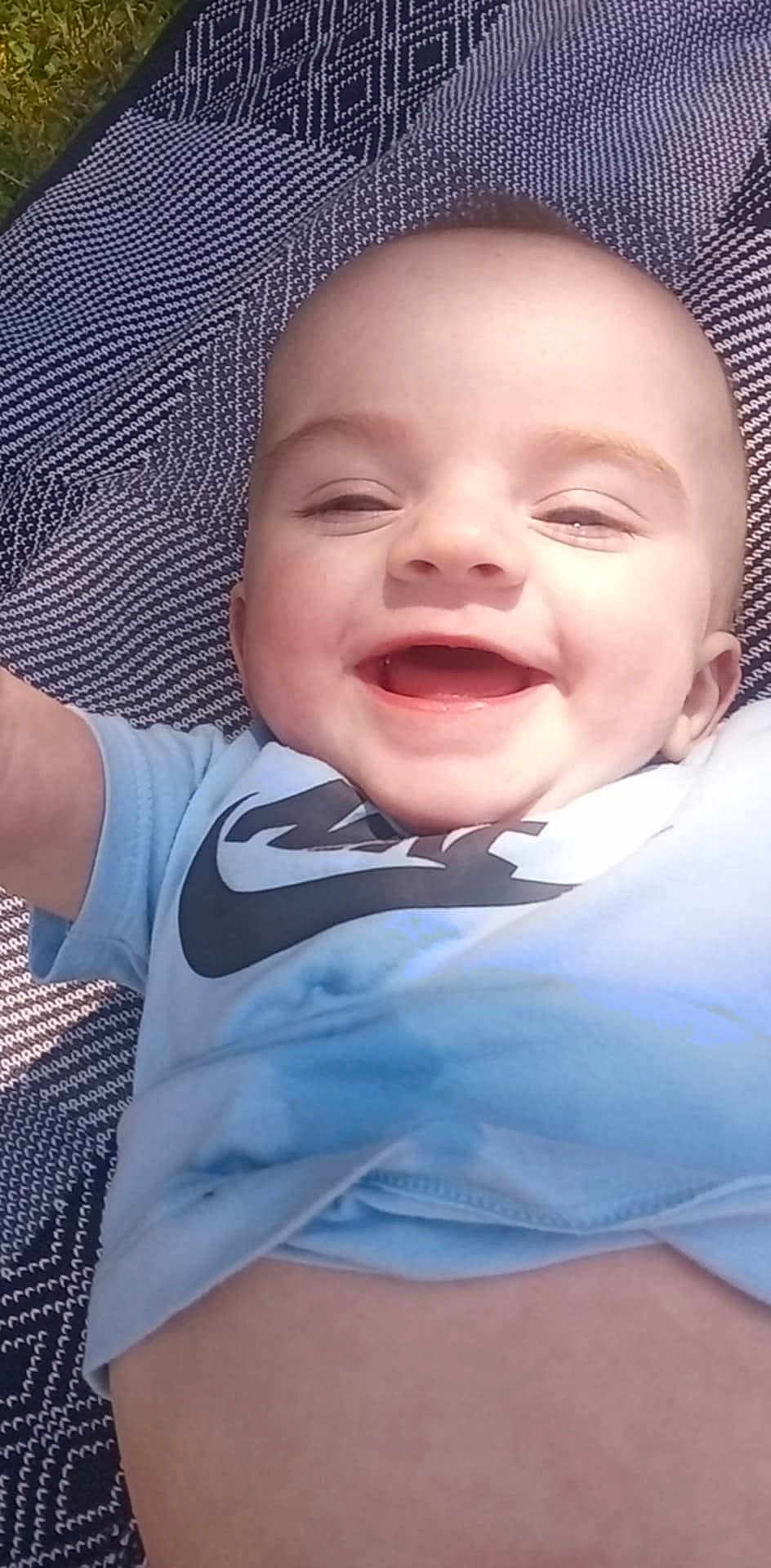 Mason is registered to the contest to win money with this photo: baby, smiling, child, outdoor, blanket, blue_shirt, face, happy, infant, toddler, pattern, grass, sunlight, portrait, cute, closeup, cheerful, clothing, skin, baby_girl