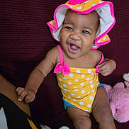A'Maya joined the competition — help win amazing prizes! baby, smiling, child, polka_dot, yellow, pink, hat, swimsuit, sitting, couch, plush_toy, indoor, happy, infant, person, clothing, headwear, cute, joyful, skin