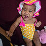 baby, smiling, child, polka_dot, yellow, pink, hat, swimsuit, sitting, couch, plush_toy, indoor, happy, infant, person, clothing, headwear, cute, joyful, skin