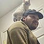 kitten, cat, man, shoulder, hat, cap, indoor, door, face, fur, curious, pet, animal, portrait, young, beard, smile, clothing, casual, light
