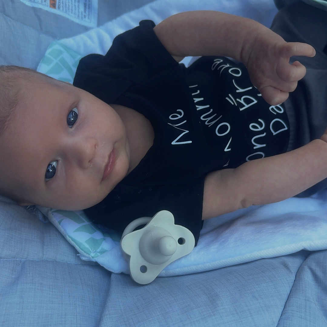 Cylan joined the competition — help win amazing prizes! baby, infant, baby_seat, pacifier, black_clothing, outdoor, cushion, face, person, child, onesie, fabric, eyes, skin, head, hand, comfort, relaxing, daylight, portrait
