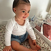 Emie a rejoint le concours — aidez-le/la à gagner de superbes lots ! toddler, child, baby, smiling, sitting, indoor, hair_buns, white_shirt, denim_shorts, barefoot, bedspread, floral_pattern, baby_products, curious, cute, person, young_child, happy, room, furniture