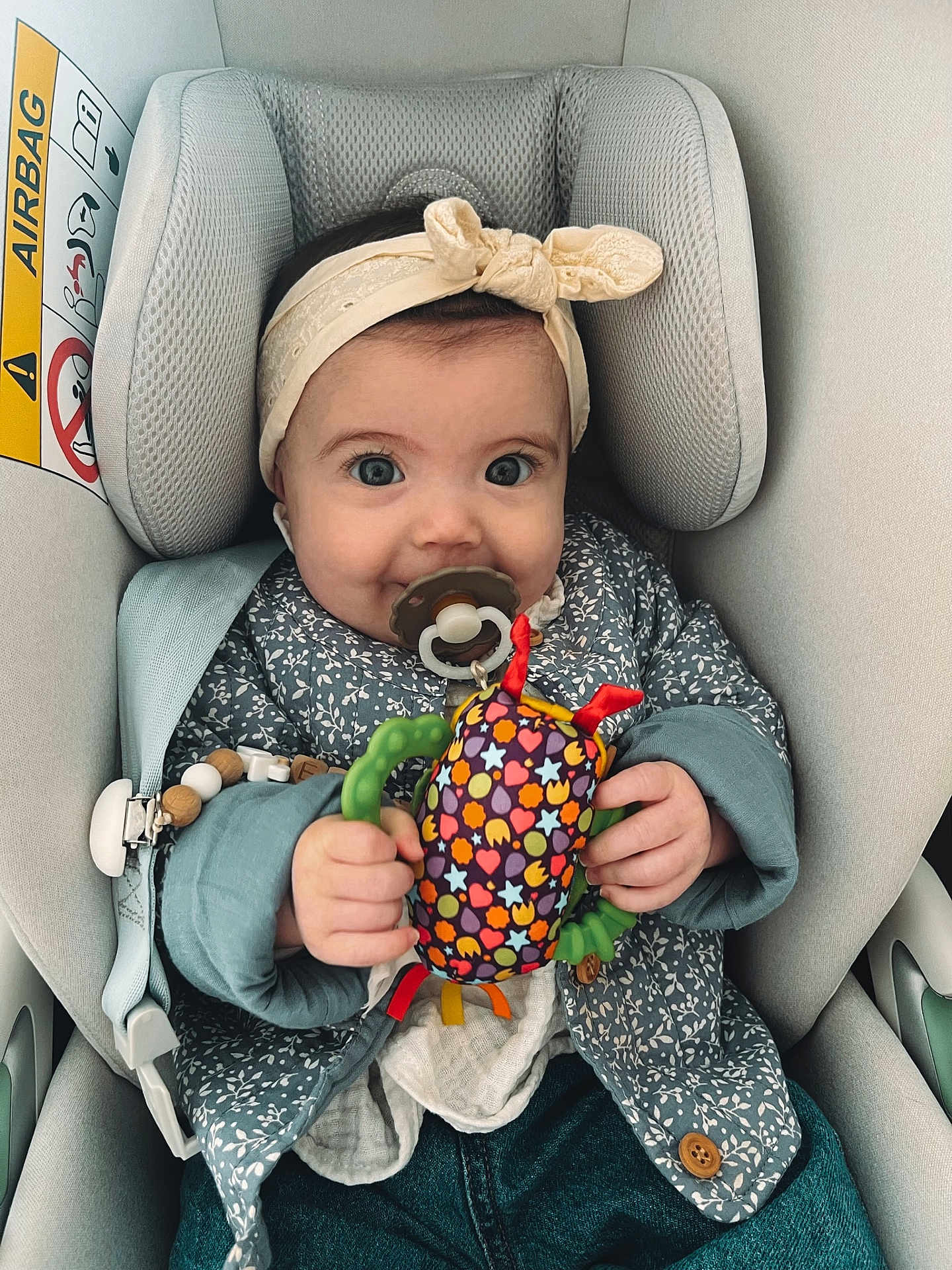 Emie a rejoint le concours — aidez-le/la à gagner de superbes lots ! baby, child, car_seat, headband, toy, pacifier, clothing, jacket, hand, face, eyes, seatbelt, indoor, comfort, cute, infant, fabric, colorful, sitting, smile