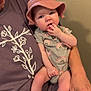 adult, baby, child, closeup, clothing, curious_expression, fingers, floral_design, glasses, hand, hat, indoor, infant, lap, onesie, person, purple_shirt, sitting, skin, wall