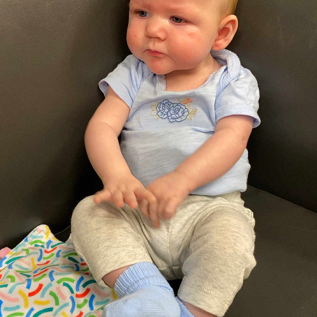 Tilly-May is registered to the contest to win money with this photo: baby, blanket, blue_clothing, child, colorful, couch, cute, expression, face, footwear, gray_pants, hands, head, indoor, infant, person, sitting, socks, toy, young_child