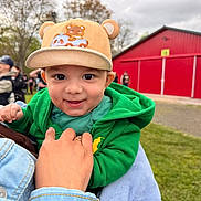 Theodore joined the competition — help win amazing prizes! baby, child, hat, cap, green_hoodie, smiling, face, person, hand, outdoor, grass, barn, red_building, cloudy_sky, trees, holding, joyful, cute, portrait, casual_clothing