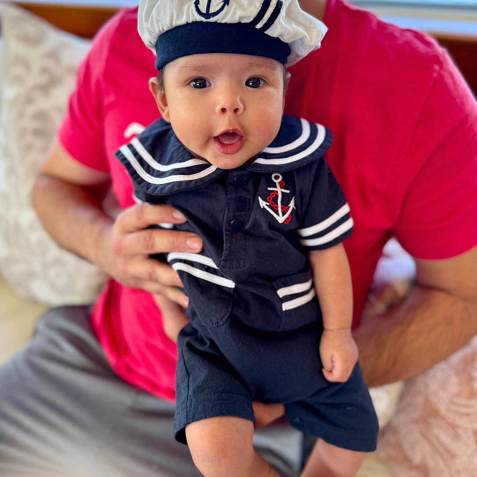 Liam joined the competition — help win amazing prizes! adult_hand, baby, child, couch, cute, eyes, face, hat, holding, indoor, infant, mouth_open, navy_blue, person, pillow, portrait, red_shirt, sailor_outfit, seated, smiling