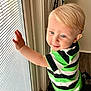 Camden is registered to the contest to win money with this photo: toddler, child, blonde_hair, blue_eyes, window, blinds, striped_shirt, green, black, white, hand, curious, indoor, natural_light, portrait, young_child, smiling, face, person, candid