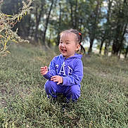 алия is registered to the contest to win money with this photo: child, toddler, purple_hoodie, outdoor, grass, nature, smile, laughing, pigtails, greenery, trees, daylight, casual_clothing, happy, cute, young, person, squatting, field, sunlight