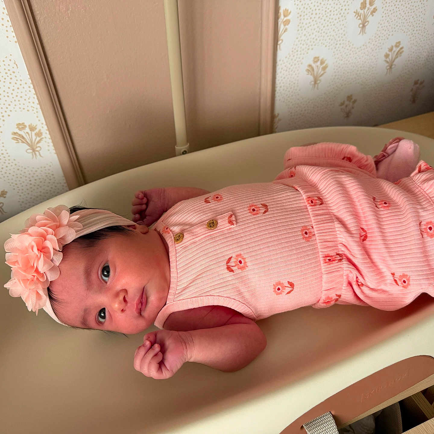Ashley is registered to the contest to win money with this photo: newborn, baby, infant, child, portrait, lying_down, pink_outfit, floral_clothing, headband, flower, changing_table, nursery, nursery_decor, wallpaper, woven_basket, indoor, facial_expression, blue_eyes, hands, safe
