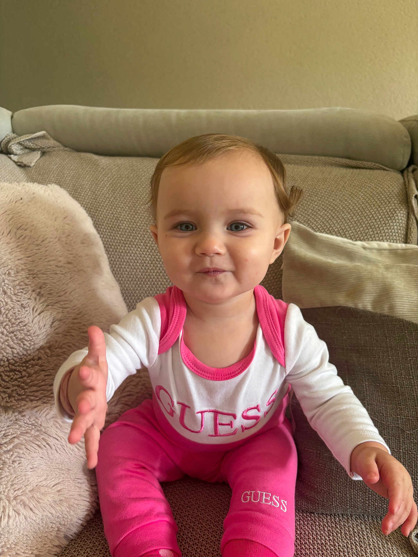 Ellie participe au concours pour gagner de l'argent avec cette photo : toddler, child, baby, pink_clothing, white_clothing, guess_brand, couch, pillow, blanket, indoor, smiling, curious, reaching_hand, sitting, portrait, cute, young_child, cozy, home, soft_texture