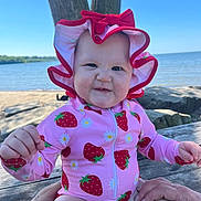 Selena is registered to the contest to win money with this photo: baby, child, swimsuit, sun_hat, strawberry_pattern, pink_clothing, outdoor, lake, tree, summer, cute, portrait, infant, hands, wooden_bench, nature, blue_sky, smile, expression, sitting