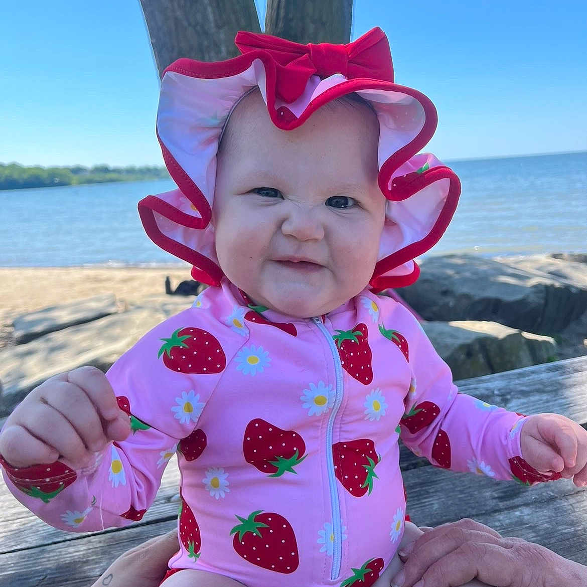 Selena is registered to the contest to win money with this photo: baby, child, swimsuit, sun_hat, strawberry_pattern, pink_clothing, outdoor, lake, tree, summer, cute, portrait, infant, hands, wooden_bench, nature, blue_sky, smile, expression, sitting