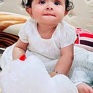 Anika joined the competition — help win amazing prizes! baby, child, white_dress, stuffed_toy, plush, curly_hair, indoors, bed, cushion, cute, innocent, portrait, smiling, person, soft_toy, happy, young_child, face, sitting, cozy