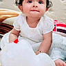 baby, child, white_dress, stuffed_toy, plush, curly_hair, indoors, bed, cushion, cute, innocent, portrait, smiling, person, soft_toy, happy, young_child, face, sitting, cozy