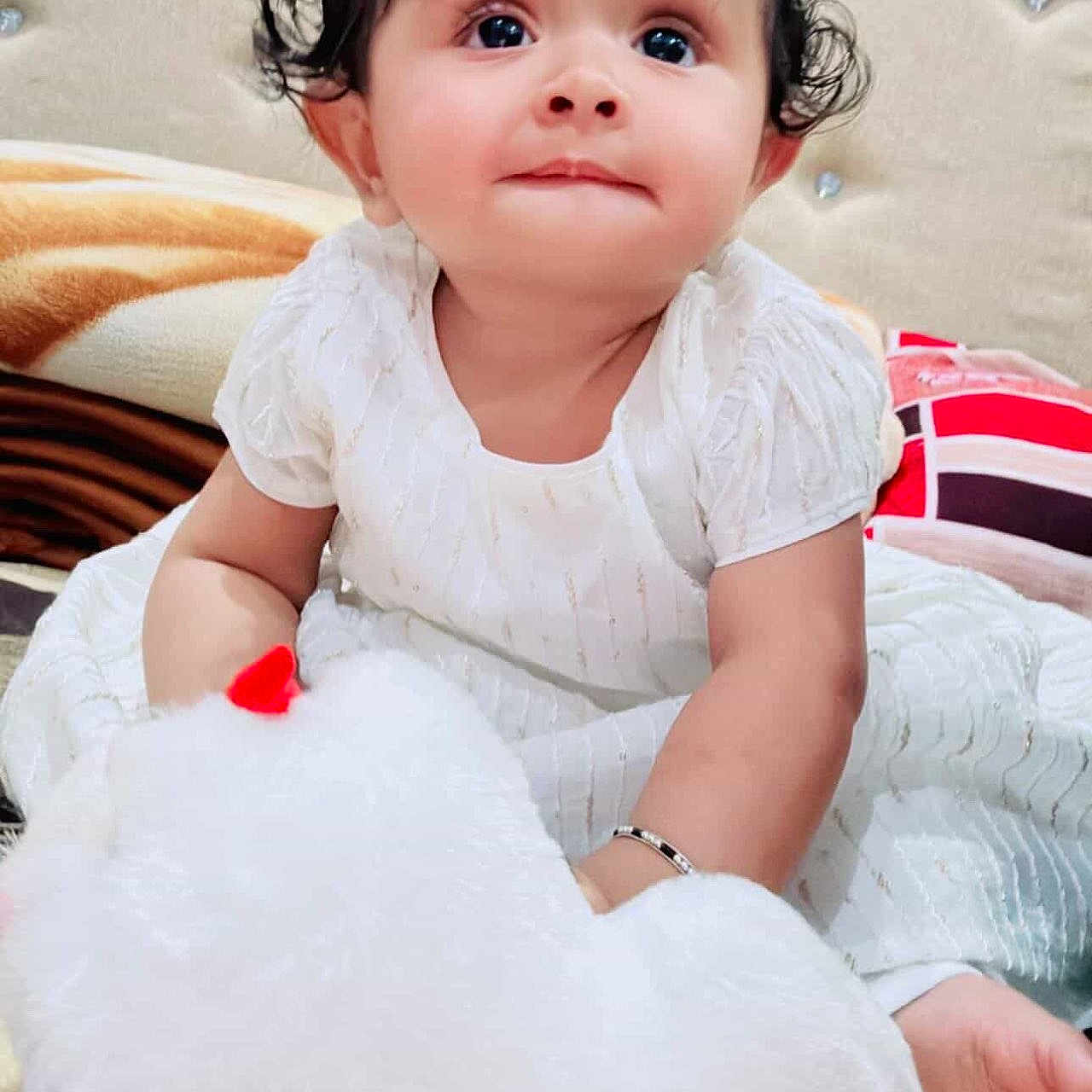 Anika joined the competition — help win amazing prizes! baby, child, white_dress, stuffed_toy, plush, curly_hair, indoors, bed, cushion, cute, innocent, portrait, smiling, person, soft_toy, happy, young_child, face, sitting, cozy