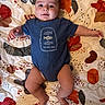 arms, baby, blue_clothing, child, colorful, cute, expression, fabric, face, head, indoor, infant, legs, lying_down, onesie, patterned_quilt, person, playful, skin, smiling