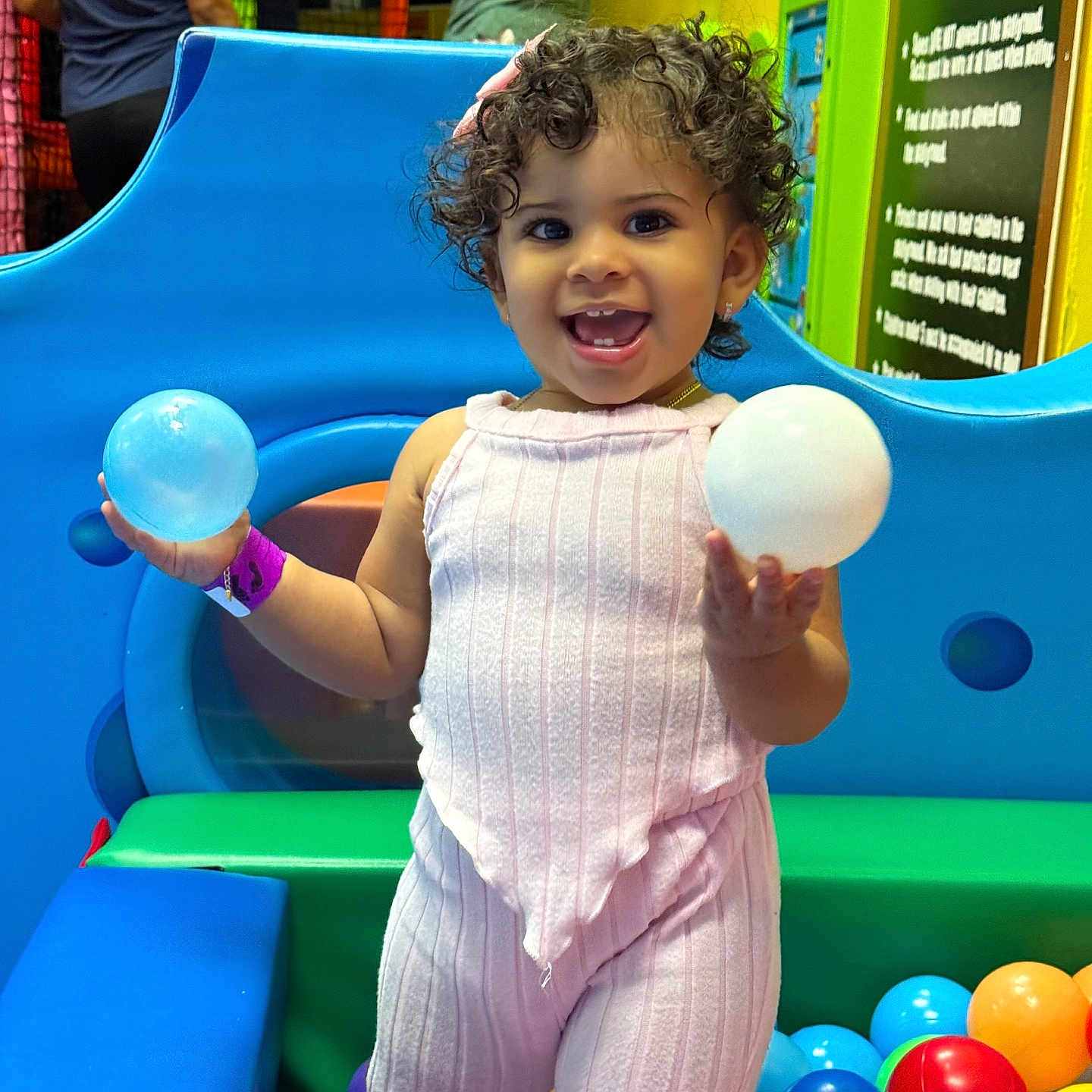 Paola is registered to the contest to win money with this photo: background_people, ball_pit, blue_bench, bright_colors, child, colorful_balls, curly_hair, earrings, green_cushion, happy, indoor_play_area, person, pink_outfit, plastic_balls, playful, playground, smiling, toddler, toy, wristband