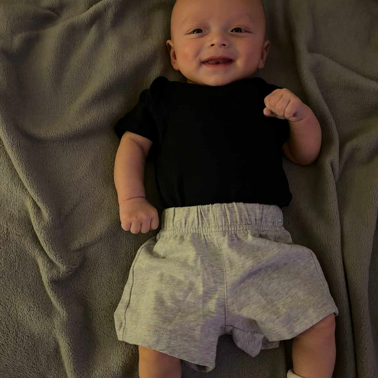 Xzavier joined the competition — help win amazing prizes! baby, child, infant, smiling, portrait, lying_down, blanket, gray_blanket, black_shirt, shorts, socks, clothing, happy, face, hands, arms, legs, indoors, cozy, photography