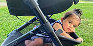 Zyon Levo joined the competition — help win amazing prizes! toddler, child, stroller, mesh_basket, grass, outdoor, curious, baby, person, wheel, black_frame, clothing, blue_stripes, hair_tie, playful, sunlight, daytime, park, resting, face