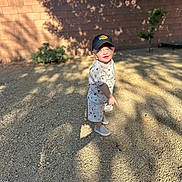 Haze is registered to the contest to win money with this photo: toddler, child, outdoor, gravel, sunlight, shadow, cap, white_clothing, shorts, sneakers, brick_wall, plant, curious_expression, standing, daylight, casual, nature_shadow, young_child, headwear, bare_arms