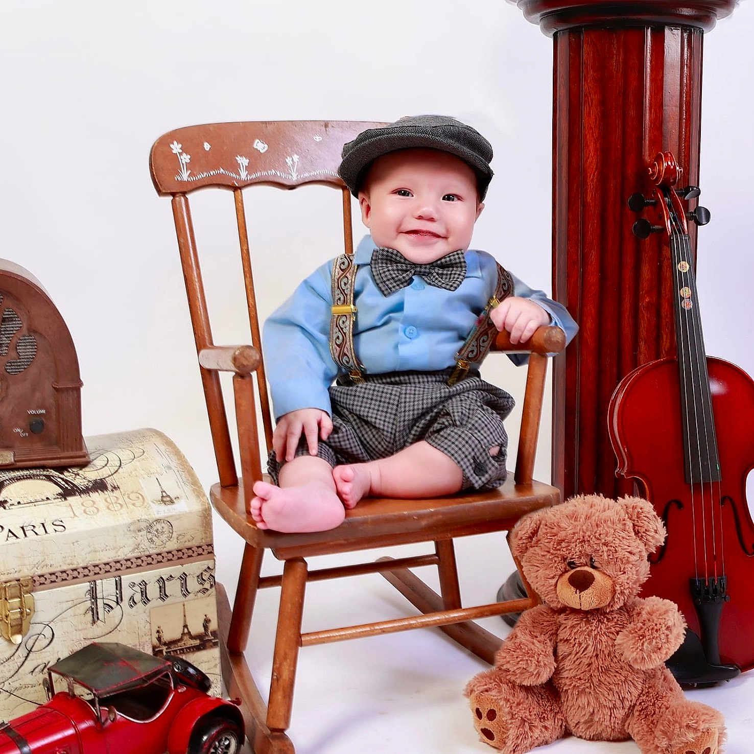 Josias is registered to the contest to win money with this photo: baby, child, vintage_clothing, bow_tie, suspenders, wooden_chair, teddy_bear, violin, toy_car, old_radio, smiling, indoor, portrait, cute, hat, props, classic_style, studio_photo, happy, person