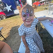 Aubrey joined the competition — help win amazing prizes! child, face_paint, unicorn, smiling, outdoor, picnic_table, food_truck, grass, sunny, dress, pigtails, happy, event, table, bench, person, festival, casual_clothing, daytime, playful