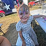child, face_paint, unicorn, smiling, outdoor, picnic_table, food_truck, grass, sunny, dress, pigtails, happy, event, table, bench, person, festival, casual_clothing, daytime, playful