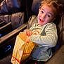 child, popcorn, movie_theater, seat, leather, smiling, happy, casual_clothing, blue_shirt, black_pants, indoor, entertainment, snack, young, person, light, hand, hair, face, fun