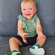Xavier is registered to the contest to win money with this photo: baby, child, smiling, sitting, couch, green_hoodie, black_shorts, sneakers, happy, blue_eyes, clapping, indoor, portrait, cute, infant, light_hair, casual_clothing, footwear, person, young