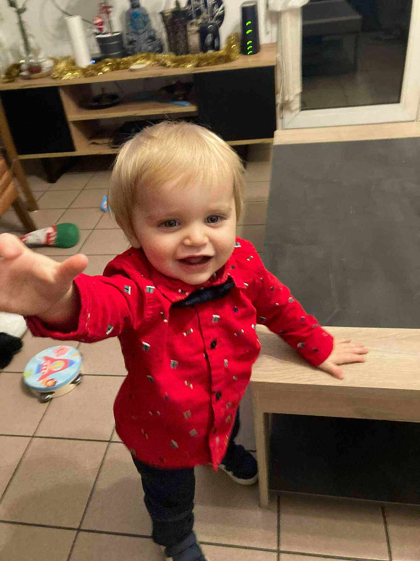 Elyo a rejoint le concours — aidez-le/la à gagner de superbes lots ! toddler, child, red_shirt, indoor, toy, floor, table, window, smiling, person, blond_hair, room, furniture, playful, young_child, casual_clothing, home, floor_tile, reaching_hand, curious