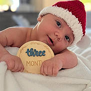 Mario is registered to the contest to win money with this photo: baby, infant, child, santa_hat, red_hat, white_blanket, soft_texture, wooden_sign, three_months, cute, portrait, indoor, close_up, smiling, holiday, christmas, festive, face, hands, lying_down