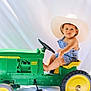 toddler, child, toy_tractor, john_deere, cowboy_hat, overalls, blue_clothing, green_toy, yellow_wheels, barefoot, cute, portrait, studio_shot, white_backdrop, sitting, playtime, outdoor_toy, young_child, curious_expression, baby