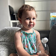 Mia joined the competition — help win amazing prizes! toddler, child, smiling, sitting, blanket, water_bottle, cotton_ball, indoor, furniture, dress, patterned_clothing, face, person, cute, baby, room, soft_light, hair, expression, comfort