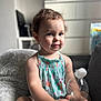 toddler, child, smiling, sitting, blanket, water_bottle, cotton_ball, indoor, furniture, dress, patterned_clothing, face, person, cute, baby, room, soft_light, hair, expression, comfort