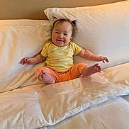 Keanna is registered to the contest to win money with this photo: baby, child, smile, bed, pillow, yellow_shirt, orange_pants, hair_ties, indoors, happy, person, cute, sitting, white_bed, cozy, comfort, portrait, young_child, room, wall