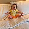 baby, child, smile, bed, pillow, yellow_shirt, orange_pants, hair_ties, indoors, happy, person, cute, sitting, white_bed, cozy, comfort, portrait, young_child, room, wall