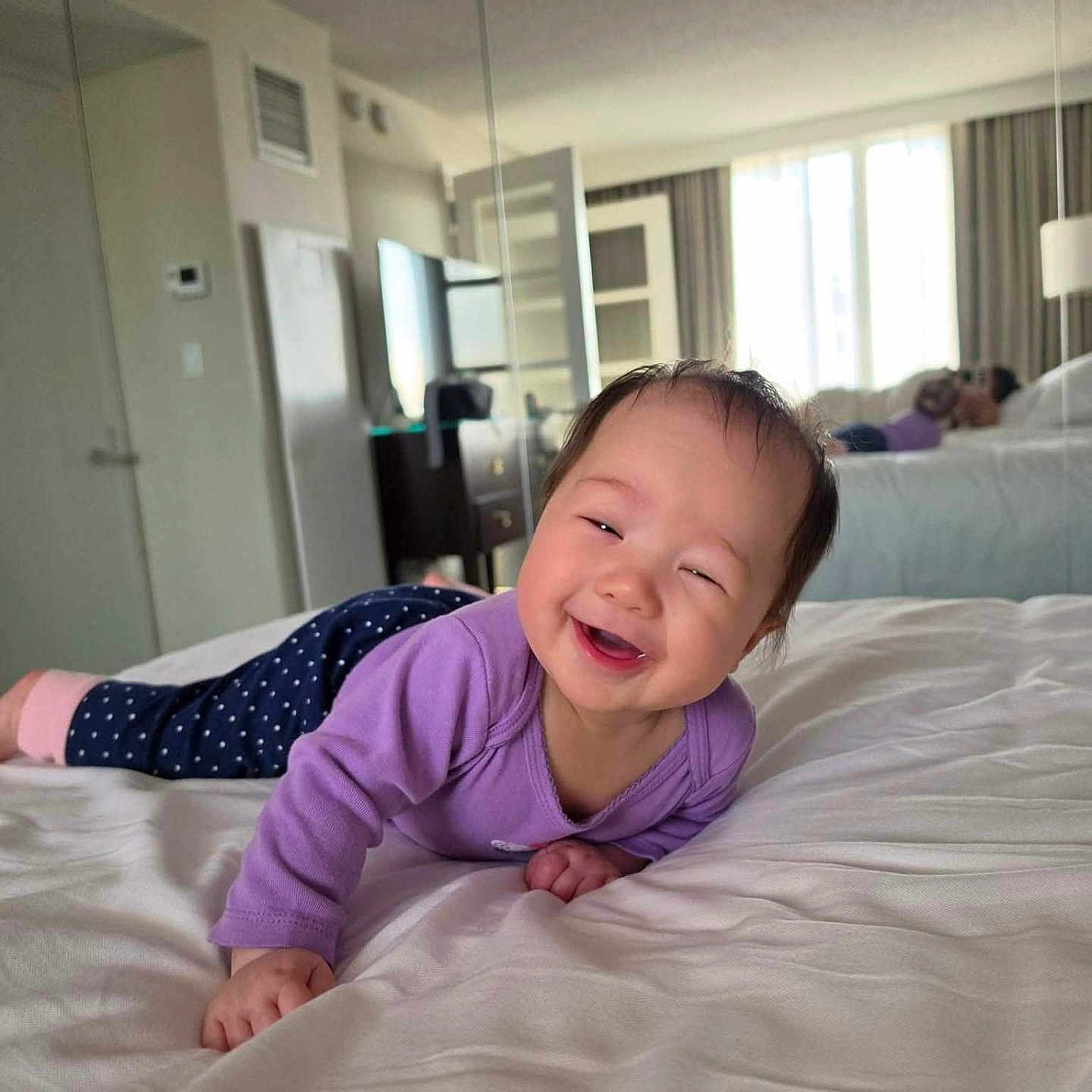 Keanna is registered to the contest to win money with this photo: baby, smiling, bed, purple_clothing, polka_dot_pants, indoor, mirror, reflection, person, furniture, window, curtains, room, happy, child, lying_down, adult, relaxing, natural_light, cozy
