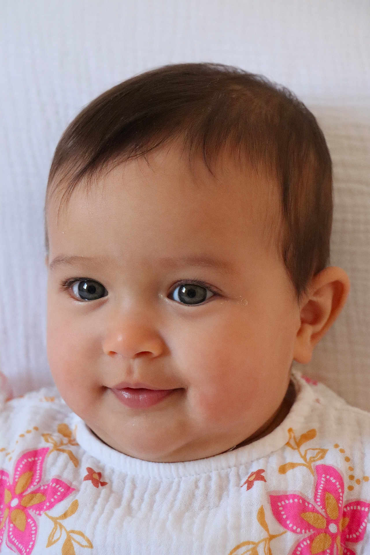 Elya participe au concours pour gagner de l'argent avec cette photo : baby, portrait, close_up, face, child, infant, smile, curly_hair, cute, eyes, clothing, pattern, floral, white_background, skin, cheeks, young, person, head, indoors