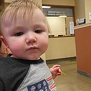 Camden is registered to the contest to win money with this photo: toddler, child, indoor, portrait, human_services, government_office, casual_clothing, baseball, curious, blond_hair, floor, wall, sign, hand, young_child, person, face, short_hair, expression, public_building