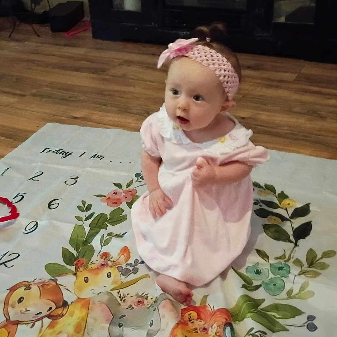 Nova is registered to the contest to win money with this photo: animal_illustrations, baby, camera, child, cute, decor, floor, hand, headband, indoor, infant, looking, milestone_blanket, months, numbers, person, photography, pink_dress, sitting, wooden_floor