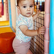 Alessia participe au concours pour gagner de l'argent avec cette photo : toddler, child, baby, playpen, netting, pink_pants, floral_shirt, bow, indoor, playroom, curious, standing, soft_padding, colorful, young_child, cute, expression, person, portrait, hand