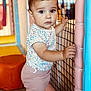 toddler, child, baby, playpen, netting, pink_pants, floral_shirt, bow, indoor, playroom, curious, standing, soft_padding, colorful, young_child, cute, expression, person, portrait, hand