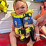 baby, barefoot, blue, child, fish_pattern, hand, life_jacket, outdoor, person, playful, red_surface, safety, sitting, smiling, striped_shirt, sunlight, toddler, white_wall, window, yellow