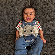 Moksha joined the competition — help win amazing prizes! baby, child, smiling, sitting, blue_overalls, elephant_patch, striped_shirt, couch, gray_fabric, barefoot, happy, indoors, portrait, cute, playful, person, infant, face, hand, foot
