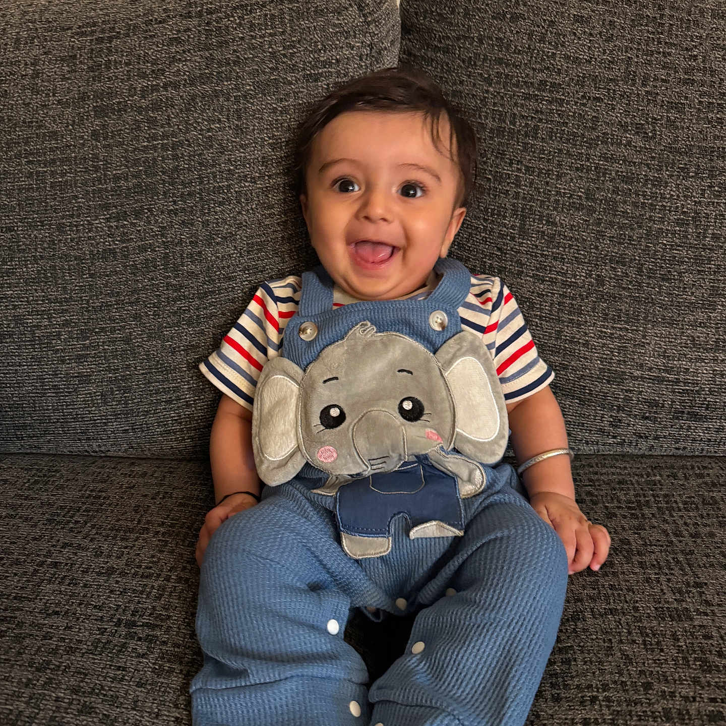 Moksha joined the competition — help win amazing prizes! baby, child, smiling, sitting, blue_overalls, elephant_patch, striped_shirt, couch, gray_fabric, barefoot, happy, indoors, portrait, cute, playful, person, infant, face, hand, foot