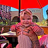baby, smile, headband, tattoo, hands, red_umbrella, outdoor, market, sunlight, table, chair, person, plaid_clothing, child, portrait, happy, face, background, daylight, holding