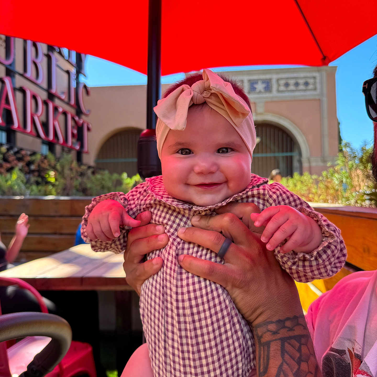 Evelina is registered to the contest to win money with this photo: baby, smile, headband, tattoo, hands, red_umbrella, outdoor, market, sunlight, table, chair, person, plaid_clothing, child, portrait, happy, face, background, daylight, holding