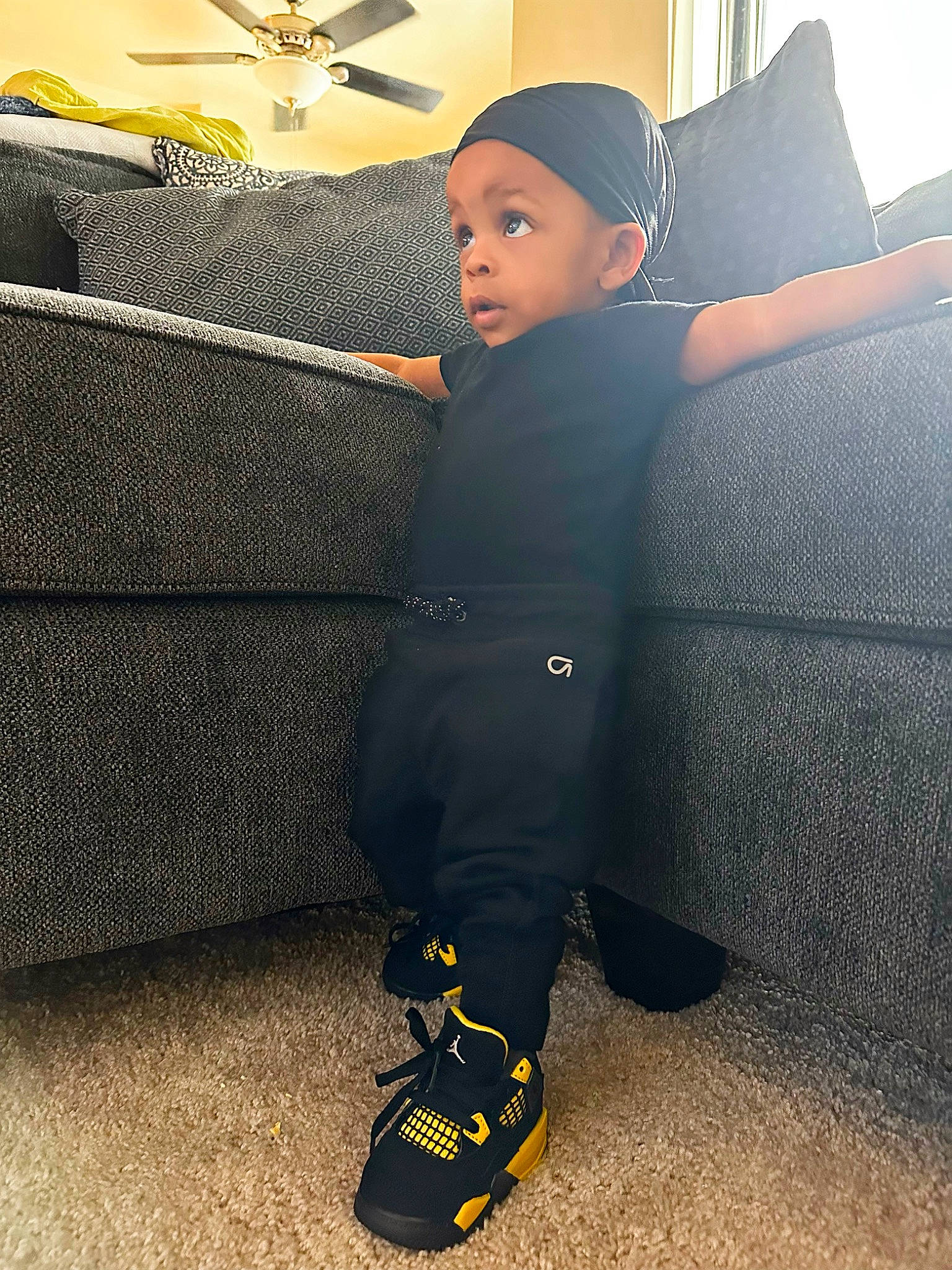 Kyron is registered to the contest to win money with this photo: baseball_cap, cap, ceiling_fan, comfort, couch, electric_blue, floor, flooring, footwear, grey, headwear, human_leg, knee, person, room, sportswear, standing, surprise, tints_and_shades, toddler