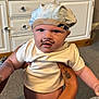 baby, infant, child, hat, white_shirt, tattoo, hand, face, mustache_drawing, indoor, carpet, cabinet, drawer_knobs, person, cute, adorable, holding, skin, expression, closeup